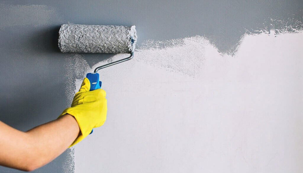 Hand in yellow glove painting wall in gray color with a roller brush.